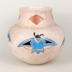 Puede incluir: Un jarrón de cerámica blanca con una superficie texturizada y un diseño geométrico. El jarrón presenta un diseño de mariposa azul, rosa y negro con detalles dorados.