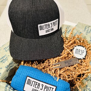 Könnte beinhalten: Eine Geschenkbox gefüllt mit braunem Seidenpapier, eine schwarz-weiße Trucker-Mütze mit dem Text "Mister 3 Putt Since 1996", ein blaues Handtuch mit dem Text "Mister 3 Putt Since 1996", ein Golfwerkzeug und ein runder schwarz-weißer Token mit dem Text "MR3P".