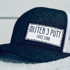 Könnte beinhalten: Eine schwarze und weiße Trucker-Kappe mit einem weißen Patch, auf dem "MJSTER 3 PUTT SINCE 1996" steht.