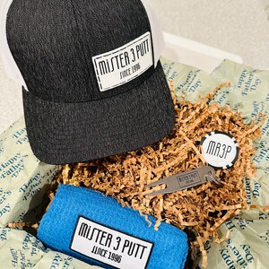 May include: A gift box filled with brown paper shred, a black and white baseball cap with a white patch that says "Mister 3 Putt Since 1996", a blue towel with a white patch that says "Mister 3 Putt Since 1996", a silver golf divot tool with a keychain that says "Mister 3 Putt Since 1996", and a silver round disc with the letters "MR3P" printed on it.