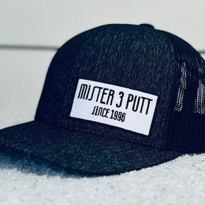 May include: A black and gray trucker hat with a white patch that says "MISTER 3 PUTT SINCE 1996".