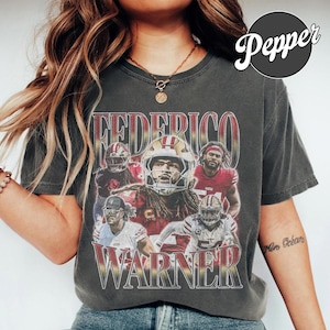 Könnte beinhalten: Ein anthrazitfarbenes Kurzarm-T-Shirt mit einem Grafikdruck des Footballspielers Fred Warner in verschiedenen Posen und Trikots. Der Text "FEDERICO" steht oben und "WARNER" unten in einer kräftigen, abgenutzten Schrift in Rot und Gold.