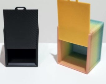 Puede incluir: Tres pequeñas cajas cuadradas con tapas. Las cajas están hechas de diferentes materiales y colores: morado, negro y un degradado arcoíris. Las tapas están abiertas en las cajas morada y arcoíris, y cerradas en la caja negra.