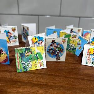 May include: Miniature Toy Story books with colorful covers. The books feature various characters and scenes from the animated movie. The books are arranged on a wooden surface, with some standing upright and others folded open.