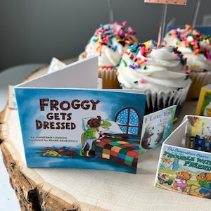 May include: A miniature book titled Froggy Gets Dressed by Jonathan London, illustrated by Frank Remkiewicz, featuring a green frog character in a bedroom. The book is displayed alongside other small books and cupcakes topped with white frosting and colorful sprinkles.