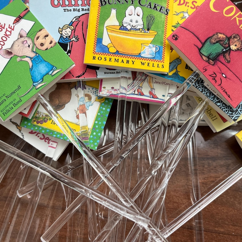 May include: A collection of miniature children's book covers featuring titles like Bunny Cakes, If You Give a Mouse a Cookie, and Corduroy, each attached to a clear, twisted acrylic rod. The colorful book covers are arranged in a layered, overlapping pile.