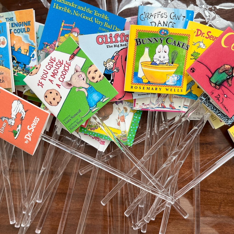 May include: A collection of children's books with colorful covers, including titles like "Bunny Cakes" and "If You Give a Mouse a Cookie." Clear plastic stir sticks are scattered throughout the scene, creating a playful arrangement.