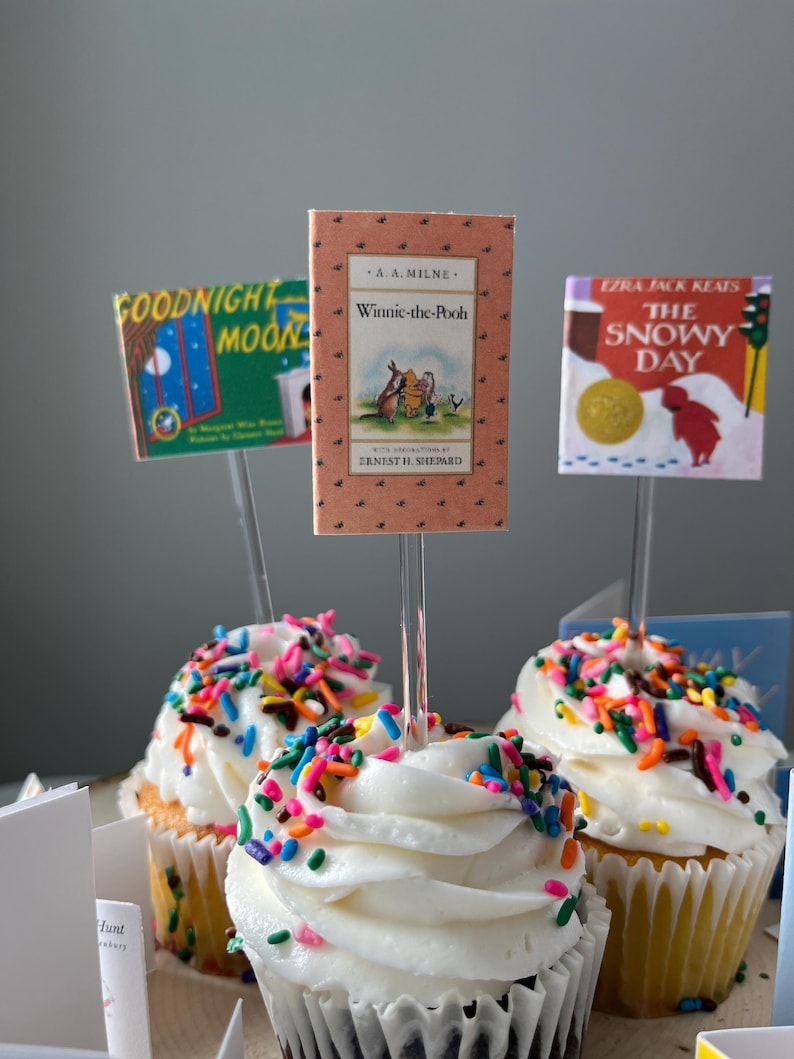Pu&ograve; includere: Tre cupcake guarniti con glassa bianca e zuccherini multicolori, ognuno dotato di un topper a forma di libro in miniatura. I topper mostrano le copertine di Goodnight Moon, Winnie-the-Pooh di A.A. Milne e The Snowy Day di Ezra Jack Keats.