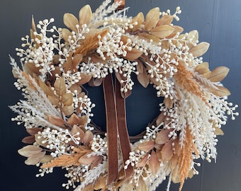 May include: A decorative wreath featuring dried leaves, white berries, and feathery grasses in shades of beige, brown, and cream. A brown velvet ribbon hangs from the top. The wreath is displayed against a dark background.