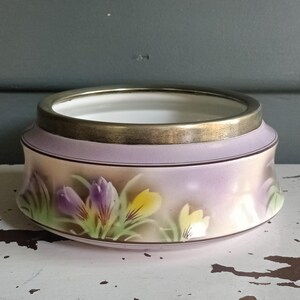 May include: A round ceramic bowl with a lavender exterior and a gold rim. The bowl features a floral design of purple, yellow, and white crocus flowers. The bowl sits on a white surface.