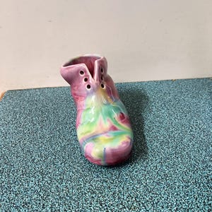 May include: A ceramic boot-shaped vase with a vibrant, swirled design. The vase features a blend of pink, green, yellow, and blue hues. It has three sets of holes on the side, and a wide opening at the top.