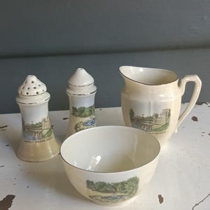 Op de afbeelding: Een set van vier witte keramische stukken met gouden rand. De stukken omvatten een roomkan, een suikerpot en twee zout- en peperstrooiers. Elk stuk heeft een ander beeld van een brug of gebouw. De roomkan heeft de tekst "The Menai Bridge" erop. De suikerpot heeft de tekst "The Menai Bridge" erop. De zoutstrooier heeft de tekst "Menai Bridge, Anglesey" erop. De peperstrooier heeft de tekst "Menai Bridge, Anglesey" erop.