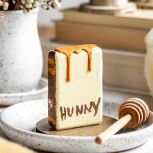 May include: A rectangular dessert with a light cream-colored body and brown edges. The top has a caramel-colored drizzle. The word "HUNNY" is written in brown on the front. A wooden honey dipper rests on a plate.