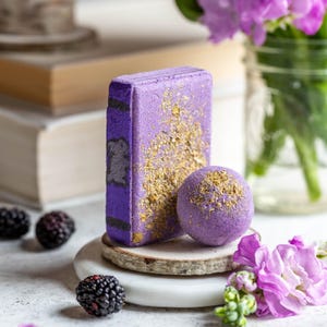 Könnte beinhalten: Lila und goldene Badebombe mit einem lila und goldenen Badeblock auf einem Holzsockel. Die Badebombe und der Badeblock befinden sich auf einer weißen Marmorfläche. Im Hintergrund sind Brombeeren und lila Blumen zu sehen.