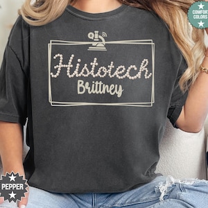 May include: Dark gray t-shirt with the text "Histotech Brittney" in a white, pearl-like font, framed by a white rectangle. A microscope graphic is above the text. The shirt has a relaxed fit and short sleeves.