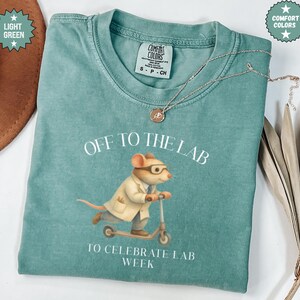 May include: A light green t-shirt with the text "OFF TO THE LAB TO CELEBRATE LAB WEEK". The shirt features a cartoon mouse wearing glasses and a lab coat, riding a scooter. A silver necklace with a pendant is on the shirt.