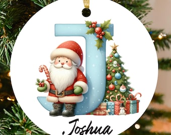 Könnte beinhalten: Runder Weihnachtsschmuck mit blauem Buchstaben "J", einer Illustration des Weihnachtsmanns und dem Namen "Joshua". Der Weihnachtsmann hält eine Zuckerstange neben einem Weihnachtsbaum und Geschenken. Dekoration hängt an einem goldenen Band.