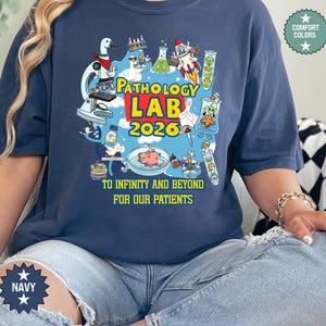 May include: Navy blue t-shirt with a colorful graphic featuring lab equipment, cartoon characters, and the text "PATHOLOGY LAB 2026." The shirt also includes the phrase "TO INFINITY AND BEYOND FOR OUR PATIENTS."