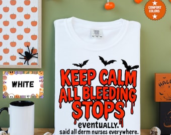 May include: White t-shirt with the text "KEEP CALM ALL BLEEDING STOPS eventually. said all derm nurses everywhere." in red. The shirt has three black bat silhouettes above the text. Halloween-themed decor surrounds the shirt.