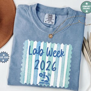 May include: A blue jean Comfort Colors t-shirt with the words "Lab Week 2026" in a blue and green design. A gold necklace with a pendant is draped across the shirt. The shirt is folded and has a small tag.