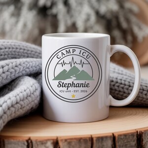 Puede incluir: Una taza de cerámica blanca con un diseño circular que incluye el texto CAMP ICU, una cordillera, una línea de latido, el nombre Stephanie y el texto ICU unit EST. 2026 con una pequeña estrella amarilla en la parte inferior.