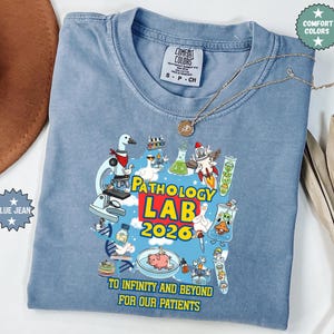 May include: Blue jean-colored t-shirt with a colorful graphic featuring lab-themed illustrations and the text "PATHOLOGY LAB 2026." The shirt also includes the phrase "TO INFINITY AND BEYOND FOR OUR PATIENTS."