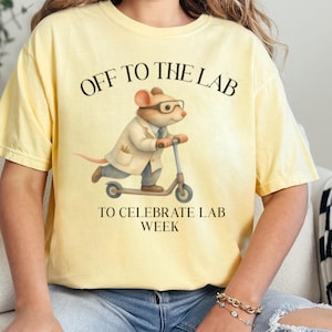 May include: Yellow t-shirt with the text "OFF TO THE LAB" and "TO CELEBRATE LAB WEEK." The graphic shows a mouse wearing glasses and a lab coat riding a scooter. The shirt is a light yellow color.