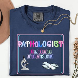 May include: Navy blue t-shirt with the words "PATHOLOGIST SLIDE READER" in a colorful, outlined font. A microscope and slides are illustrated below the text. A gold necklace with a star pendant is draped over the shirt.