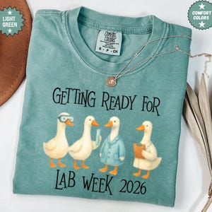 May include: A light green Comfort Colors t-shirt with the text "GETTING READY FOR LAB WEEK 2026". The shirt features a graphic of four ducks dressed as scientists. A silver necklace with a pendant is on the shirt.