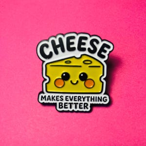 May include: A yellow enamel pin shaped like a block of Swiss cheese with a smiling face, black outlines, and the text CHEESE MAKES EVERYTHING BETTER in black capital letters. The design features two orange circles for cheeks and black eyes.