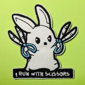 May include: A white embroidered patch featuring a cartoon rabbit with black outlines holding two pairs of blue scissors. Below the rabbit, a black rectangular banner contains the white embroidered text "I RUN WITH SCISSORS" in a bold, blocky font.