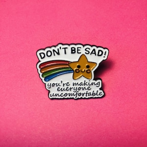 Puede incluir: Un pin de esmalte blanco con forma de bocadillo que presenta una estrella amarilla con cara sonriente, una estela de arcoíris y el texto negro DON'T BE SAD! you're making everyone uncomfortable. El pin tiene un acabado brillante y bordes negros.