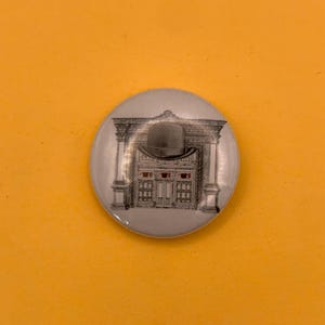 Puede incluir: Un pin redondo con una ilustración en blanco y negro de un arco arquitectónico con un bombín sobre la entrada de una tienda. El escaparate muestra una hilera de ojos humanos en los cristales superiores de la ventana.