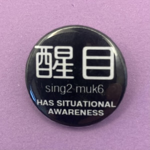 May include: A circular black pin-back button featuring white text. The top displays two large Chinese characters, followed by the text "sing2 muk6" in the middle, and the phrase "HAS SITUATIONAL AWARENESS" printed in capital letters at the bottom.