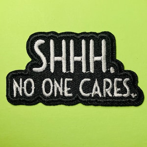Könnte beinhalten: Ein schwarzer bestickter Aufnäher mit gewelltem Rand und dem in Weiß gestickten Text SHHH. NO ONE CARES. Das Design ist in zwei Zeilen angeordnet, wobei das Wort SHHH. oben und NO ONE CARES. unten in einer fetten Schriftart steht.
