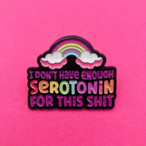 Peut inclure: Une broche en émail noir représentant un arc-en-ciel coloré au-dessus de deux nuages blancs. Le texte indique I DON'T HAVE ENOUGH SEROTONIN FOR THIS SHIT en lettres multicolores. La broche présente un fond sombre et un design audacieux.
