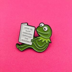 May include: A green frog enamel pin featuring a white book held by the frog. The book cover has black text that reads: HOW TO SLAP SOMEONE THROUGH THE INTERNET. The pin has a black metal outline and a smooth, glossy finish on the frog and book.