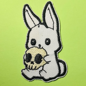 May include: An embroidered patch featuring a white rabbit with black outlines holding a pale yellow skull. The rabbit has large, dark eyes and long ears, while the skull has deep black eye sockets and a detailed jawline, all bordered by black thread.