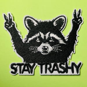 May include: An embroidered patch featuring a black and white raccoon with its arms raised in peace signs. The raccoon has a white face mask and chest fur. Below the raccoon, the text STAY TRASHY is embroidered in bold black capital letters.