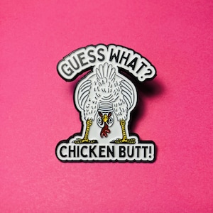 May include: A white enamel pin featuring a chicken bending over with its head between its legs. The text GUESS WHAT? is arched above the chicken, and the text CHICKEN BUTT! is printed below. The chicken has yellow legs and a red wattle.