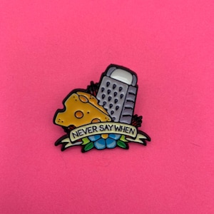 May include: A metal enamel pin featuring a yellow block of Swiss cheese with holes next to a grey metal cheese grater. Below the items is a cream-coloured banner with black text that reads NEVER SAY WHEN, accented by a blue and green flower.