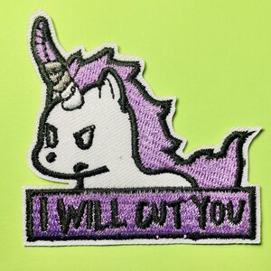 May include: An embroidered patch featuring a white unicorn with a purple mane and a sharp horn. The unicorn has an angry expression and is positioned above a purple rectangular banner with the black text I WILL CUT YOU.