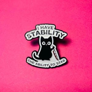 May include: A black and white enamel pin featuring a cartoon cat holding a knife. The text above the cat reads I HAVE STABILITY and the text below reads THE ABILITY TO STAB. The design has a black outline and white background elements.