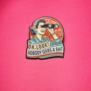 Peut inclure: Un pin's en émail circulaire représentant une illustration rétro d'une personne portant un foulard et regardant à travers des jumelles noires. Le fond est un cercle corail avec des étoiles. Le texte indique : OH, LOOK! NOBODY GIVES A SHIT.