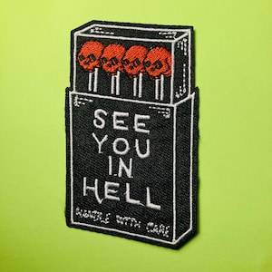 Puede incluir: Un parche bordado rectangular con un diseño de caja de cerillas negra y contornos blancos. La caja contiene cuatro cerillas con cabezas naranjas en forma de calavera. El texto dice SEE YOU IN HELL y HANDLE WITH CARE en letras blancas.
