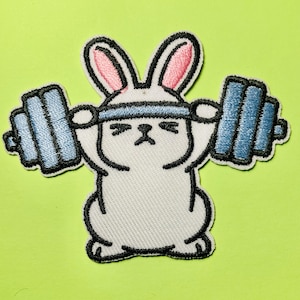 May include: An embroidered patch featuring a white rabbit with pink-lined ears lifting a blue barbell over its head. The rabbit has a determined facial expression with closed eyes and a small nose, outlined in black thread on a white background.