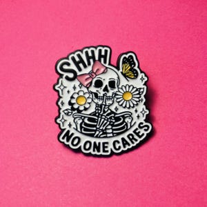 Peut inclure: Une broche en émail blanc représentant un squelette noir avec un doigt sur la bouche, un nœud rose sur la tête, deux marguerites blanches et un papillon jaune. Le texte SHHH et NO ONE CARES est écrit en lettres capitales noires autour du motif.