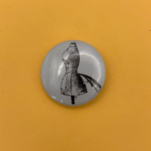 Peut inclure: Un badge rond avec un fond gris clair et une illustration graphique en noir et blanc représentant un mannequin de couture. Le mannequin est représenté dans un style de dessin à l'encre légèrement vieilli avec un pied de support.