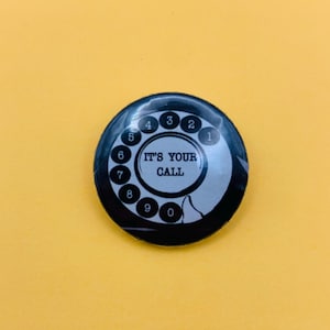 Puede incluir: Una chapa redonda con un gráfico en blanco y negro de un dial de teléfono de disco. En el centro del dial aparece el texto IT'S YOUR CALL en letras mayúsculas negras, rodeado por los números del uno al cero dispuestos en un círculo.