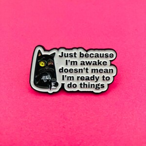 Peut inclure: Une broche en émail noir représentant un chat noir stylisé avec un œil jaune et un œil fermé, tenant une tasse à café blanche. La broche porte le texte blanc : Just because I'm awake doesn't mean I'm ready to do things, sur fond blanc.